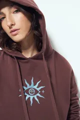 Canguro negro de calce relajado con capucha y cordones ajustables. Presenta un estampado gráfico frontal de un ojo con rayos de sol y la frase "STAY WEIRD".