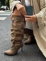 Botas de caña alta color camel, confeccionadas en material tipo gamuza, con diseño slouchy y puntera fina. Presentan dos correas con hebillas metálicas, una en la parte superior de la caña y otra en el tobillo, y cuentan con un taco fino.