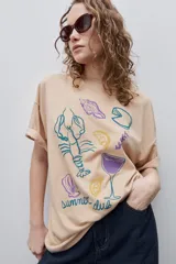 Remera color tostado de corte holgado, con cuello redondo y mangas cortas. Presenta un estampado gráfico frontal de estilo veraniego con ilustraciones de una langosta, conchas, limón, una copa y la inscripción 'Summer Club'.