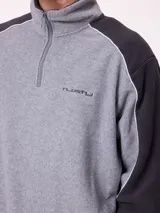 Buzo de polar con cierre frontal parcial, cuello alto y diseño de bloques de color en gris y negro. Presenta logo bordado en el pecho y puños elásticos.