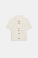 Camisa de manga corta en color crudo, con cuello solapa y cierre frontal mediante cremallera oculta. Presenta un diseño central con volantes verticales y tachas metálicas decorativas a lo largo de la botonadura.