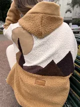 Campera con capucha de tela tipo bucle y paño, con diseño de paisaje nórdico en la espalda que combina texturas y tonos tierra. Presenta bolsillos frontales y etiqueta de cuero sintético con logo de la marca.
