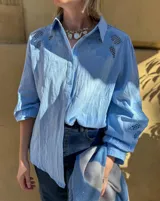 Camisa blanca de corte amplio con mangas tres cuartos abullonadas. Presenta detalles de bordado calado en los hombros y en las mangas, con cierre frontal de botones.