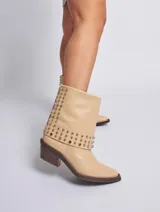 Bota de cuero color marrón con caña media, taco cuadrado y punta fina. Presenta tachas plateadas en el borde superior y en la tira que rodea el tobillo. El cuero tiene un acabado liso y brillante.