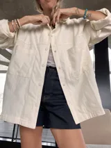 Sobre camisa color crema con capucha, cierre frontal con botones a presión, bolsillos con solapa en el pecho y mangas largas con puños abotonados.