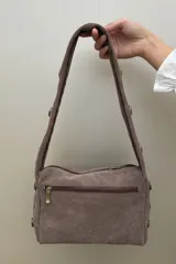 Cartera de hombro confeccionada en gamuza color habano, con diseño de correa ancha decorada con tachas metálicas circulares. Presenta cierre superior y placa metálica con logo en el frente.