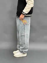 Pantalón de jean estilo baggy de corte ancho y relajado, color azul claro con lavado desgastado. Presenta un bordado en el bolsillo trasero derecho con un diseño de llamas y las siglas SDA.