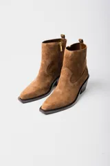 Bota corta de estilo western confeccionada en cuero gamuzado color marrón. Presenta un diseño de caña media con tirador trasero, corte asimétrico en el empeine, punta cuadrada y taco bloque de madera.