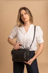 Bandolera de diseño rectangular en color suela, confeccionada en material sintético. Cuenta con un asa superior fija, correa larga ajustable y extraíble, y dos bolsillos frontales con cierre metálico, uno de ellos con diseño superpuesto. Presenta el logo de la marca grabado en el frente.