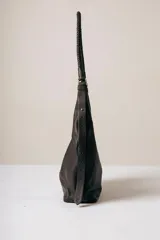 Cartera tipo hobo de cuero negro, con diseño de líneas suaves y caída natural. Presenta una manija trenzada fija y costura central vertical en el frente.