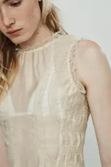 Camisa sin mangas color crema, con cuello redondo y detalle de encaje a tono en el cuello y los bordes de las sisas. Presenta un fruncido horizontal en la parte inferior y cierre con botones forrados en la espalda.
