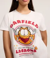 Remera de algodón blanco con cuello redondo y manga corta. Presenta un estampado frontal del personaje Garfield con cubiertos y los textos 'Garfield' y 'Lasagna' en color rojo.