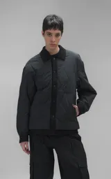 Campera tipo sobrecamisa de diseño oversize, confeccionada en microfibra opaca con patrón matelaseado geométrico. Presenta cuello, puños, cartera central y ruedo en gabardina de algodón, con cierre frontal mediante broches a presión. El diseño del ruedo simula una prenda superpuesta.
