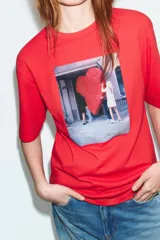 Camiseta de algodón color rojo, corte regular fit, cuello redondo y manga corta. Presenta una estampa fotográfica en el centro con dos personas sosteniendo un gran corazón rojo inflable. Incluye texto con nombre y copyright.