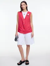 Chaleco sin mangas color coral, con escote en V, cierre frontal con botones de madera y bolsillos de parche. Incluye tres pins decorativos: un corazón plateado, un círculo plateado y un cuadrado rojo. Confeccionado en tejido liviano de lino y viscosa.