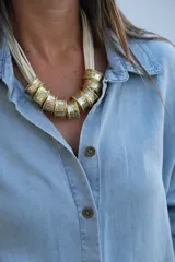 Collar de diseño artesanal compuesto por múltiples tiras de cuero en color crudo y piezas cilíndricas de metal dorado con textura martillada.
