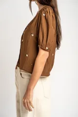 Blusa de viscosa color marrón, con escote en V, mangas cortas abullonadas y cierre frontal con botones. Cuenta con pequeños detalles bordados distribuidos por la prenda.