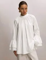 Blusa blanca de algodón con cuello alto fruncido, corte amplio y mangas largas terminadas en doble volado.