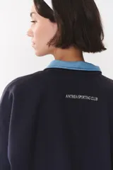 Buzo estilo rugby de corte oversized, color verde oscuro, con cuello tipo camisa en denim azul claro y cierre frontal parcial. Presenta un recorte horizontal en el pecho, logo bordado y tiras laterales en las mangas en contraste.