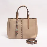 Cartera tipo tote de cuero sintético beige, con doble asa de hombro en tono tostado y correa extraíble regulable. Cuenta con detalles de costuras decorativas y un dije colgante de cordón con cuentas de colores.