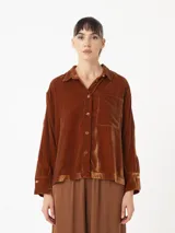 Camisa corta de terciopelo color marrón, de silueta cuadrada oversized, con bolsillo delantero y detalle de botón dorado en forma de flor en puños y cuello.