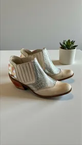 Botas cortas de cuero color natural con inserciones de cuero plateado con textura de cocodrilo en el empeine, paneles laterales elásticos y detalles pintados a mano en el talón. Cuenta con taco de madera de 5 cm.
