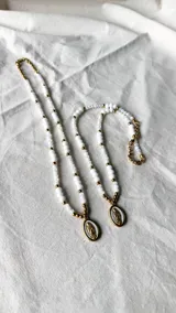 Collar largo con cuentas blancas facetadas y pequeñas esferas doradas intercaladas, con un dije central ovalado que presenta la imagen de la Virgen Milagrosa en relieve sobre fondo blanco y borde dorado.
