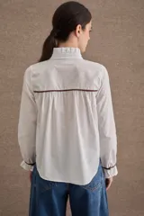 Camisa de popelina blanca con cuello alto tipo victoriano, volados frontales y detalles de costuras y ribetes en color marrón. Presenta mangas largas con puños elásticos terminados en volado y cierre frontal con botones oscuros.