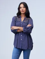 Blusa color beige de gasa con cuello a la base, escote en V y abertura central con botones. Presenta detalles de crochet en hombros y espalda, mangas 3/4 amplias con puño y botón. El largo de la espalda es mayor al delantero.