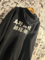 Campera negra con capucha y cierre frontal, con estampado en la espalda de texto en japonés en color blanco. Diseño inspirado en el anime Haikyu!!