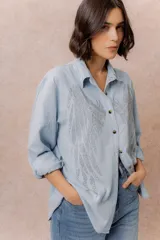 Camisa de denim liviano con calce relajado, cuello camisero clásico y cierre frontal con botones. Presenta una destacada aplicación de strass en el frente formando un diseño de alas.