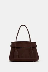 Bolso tipo citybag confeccionado en serraje color marrón oscuro. Presenta una solapa frontal con cierre magnético, asas de hombro integradas y detalles de hebillas metálicas en los laterales para ajuste. Incluye bolsillo interior con cremallera.