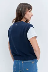 Chaleco de punto rojo con cierre frontal de botones, cuello redondo y dos bolsillos frontales con solapa. Presenta un diseño de tejido acanalado en los bordes y un pequeño parche decorativo en el pecho.