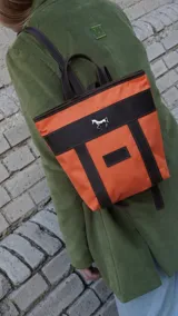 Mochila pequeña color borgoña con detalles en cuero marrón y aplique metálico de un caballo. Tiene manija superior y correas traseras regulables.