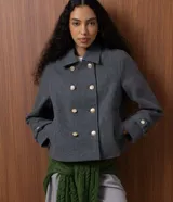 Saco corto de corte regular confeccionado en polivelour gris, con cuello inglés y cierre frontal de doble abotonadura con botones dorados. Presenta mangas largas con detalle de trabas en los puños.