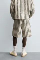 Bermuda de hombre color beige con estampado de rayas verticales azules con efecto desgastado. Corte relaxed fit, con cierre frontal de cremallera y botón, bolsillos delanteros y bolsillos traseros de plastrón.