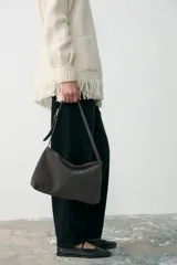 Cartera de hombro mediana confeccionada en cuero agamuzado color gris, con cierre superior y correa ajustable mediante hebilla metálica.
