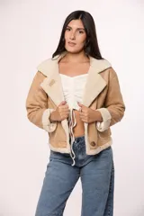 Campera tipo gamulán de gamuza color tostado con interior y solapas de corderito sintético color crema. Presenta cierre frontal con botones, bolsillos cuadrados delanteros y detalles de costuras visibles.