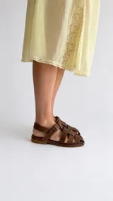 Sandalias de cuero color marrón, estilo romano con tiras entrelazadas, punta cuadrada y cierre de hebilla metálica en el tobillo.