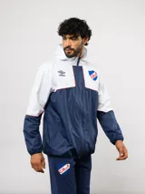 Campera de lluvia Umbro del Club Nacional de Football, con diseño de bloques de color en rojo y azul marino. Presenta capucha, cierre frontal, puños elásticos y detalles de piping blanco. Incluye logo de Umbro y escudo del club en el pecho.