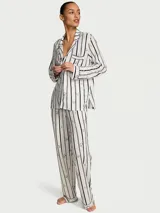 Pijama largo compuesto por una camisa de manga larga con cuello solapa, cierre frontal de botones y bolsillo en el pecho, y un pantalón de corte recto con cintura elástica. Presenta un diseño de rayas verticales oscuras sobre fondo blanco con sutiles detalles florales.