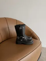 Bota de caña media estilo biker, confeccionada en eco cuero negro. Presenta suela track con plataforma, puntera redondeada y detalles de correas con hebillas metálicas y argollas decorativas en el tobillo.