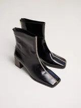 Botas de cuero color marrón con caña media y punta cuadrada. Presentan un cierre trasero, tirador para calzar fácilmente y bordado en color contrastante en el empeine. Suela de goma y taco de madera de 6 cm de altura.