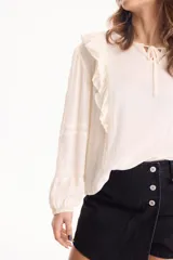 Blusa de viscosa color blanco con escote en V y cordón ajustable, mangas largas y volados decorativos en los hombros.