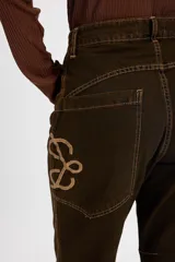 Jean tipo baggy de pierna ancha y recta, color marrón oscuro. Presenta detalles de costuras decorativas, bolsillos laterales redondeados y bolsillos traseros grandes. Incluye tiras ajustables con hebillas metálicas en los tobillos para un efecto fruncido.