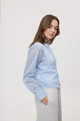 Camisa de manga larga con diseño de rayas verticales en tonos celestes, blancos y marrones, confeccionada en tejido semitransparente con cuello camisero y cierre frontal de botones.