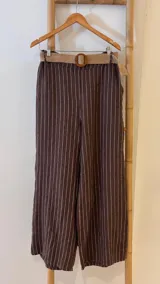 Pantalón de lino marrón con rayas verticales blancas, corte recto, cintura con elástico ancho y cinturón de rafia extraíble.