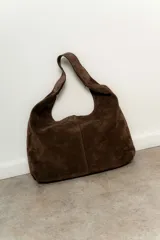 Cartera tipo tote de tamaño mediano, confeccionada en cuero gamuzado color marrón oscuro. Presenta un diseño minimalista de silueta holgada con costura central vertical y asa integrada al cuerpo de la cartera.