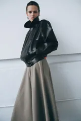 Chaqueta de estilo biker confeccionada en tejido efecto piel con acabado brillante. Presenta cuello con solapas, cierre frontal con cremallera y botones, bolsillos delanteros y bajo ajustable con trabillas y hebilla metálica.