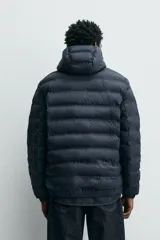 Campera puffer acolchada de corte regular fit en color negro, confeccionada en tejido técnico repelente al agua. Presenta cuello subido con capucha, cierre frontal de cremallera, mangas largas con puños elásticos y bolsillos laterales de vivo.